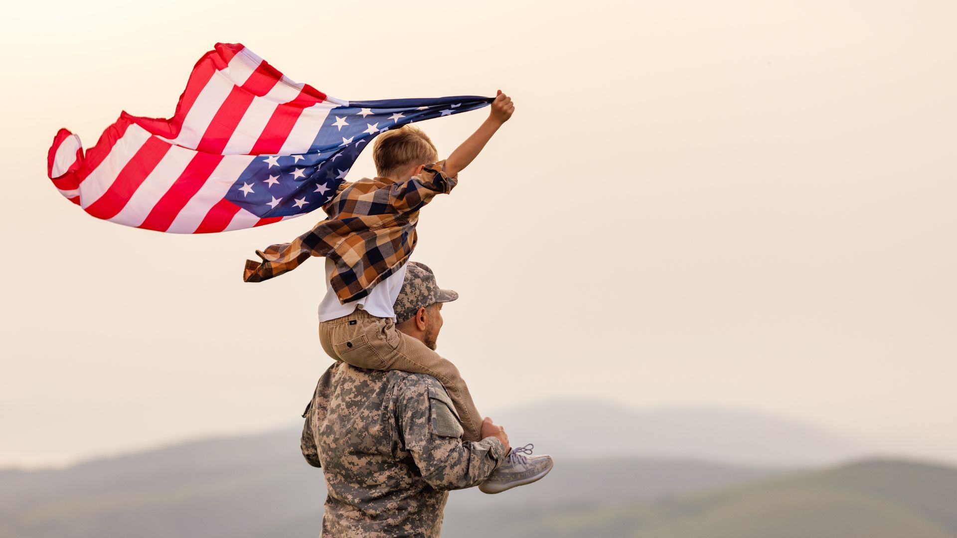 American flag being held by a child on a soldier's shoulders, symbolizing patriotism, family, and national pride. Patriotic American family scene, outdoors with mountains in the background, freedom, and national celebration.
