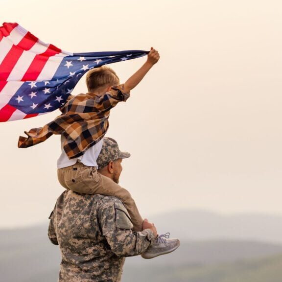 American flag being held by a child on a soldier's shoulders, symbolizing patriotism, family, and national pride. Patriotic American family scene, outdoors with mountains in the background, freedom, and national celebration.